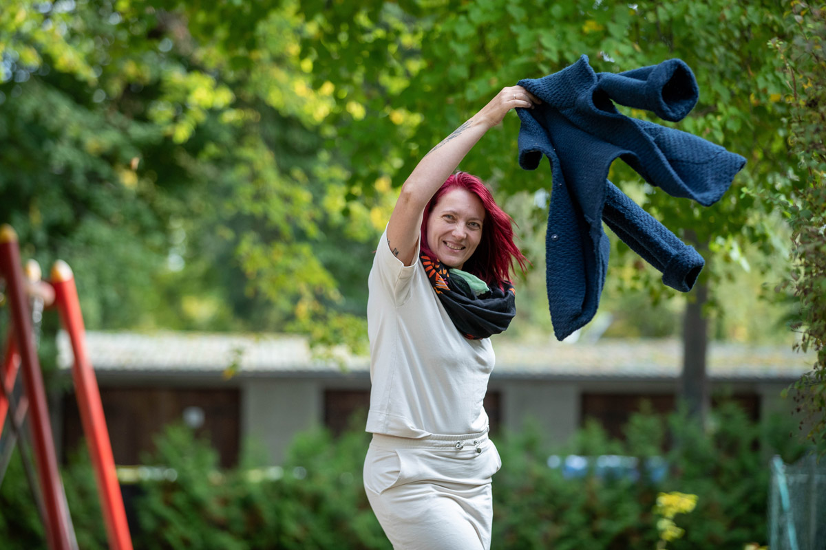 Porträt eines jungen Mannes, der an einen Rezeptions-Tresen lehnt, den rechten Ellbogen darauf aufgestützt. Er lächelt, hat dunkle Haare und trägt einen schwarzen Pullover mit hochgekrämpelten Ärmeln sowie eine helle Bundfaltenhose. In den Händen hält er einen Prospekt.