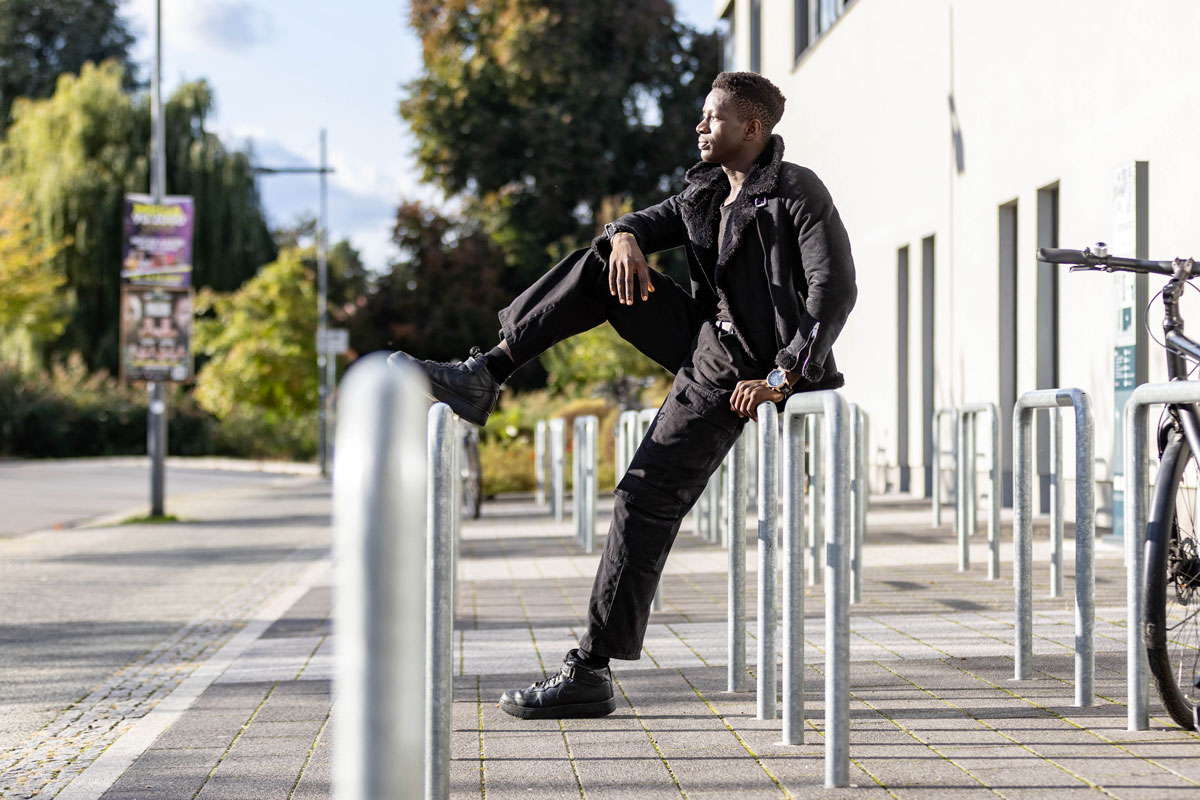 Porträt eines jungen Mannes, der an einen Rezeptions-Tresen lehnt, den rechten Ellbogen darauf aufgestützt. Er lächelt, hat dunkle Haare und trägt einen schwarzen Pullover mit hochgekrämpelten Ärmeln sowie eine helle Bundfaltenhose. In den Händen hält er einen Prospekt.