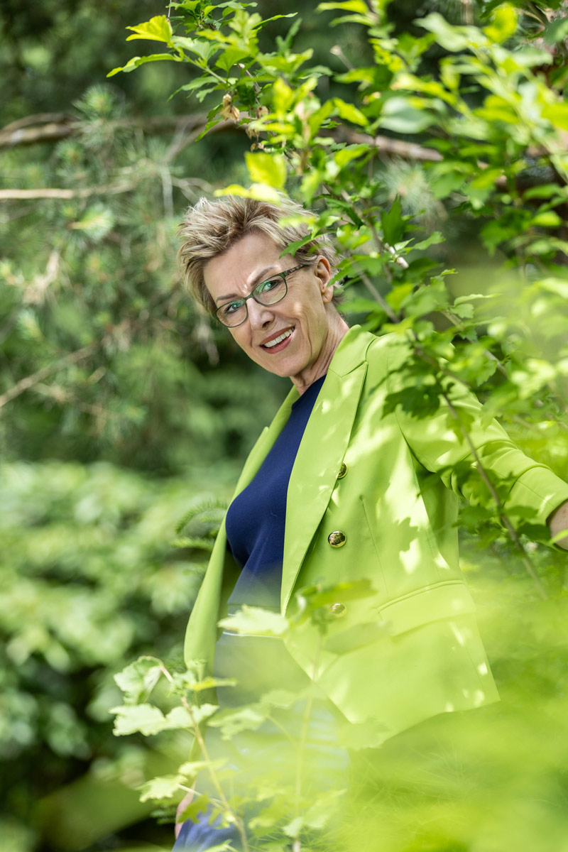 Porträt eines jungen Mannes, der an einen Rezeptions-Tresen lehnt, den rechten Ellbogen darauf aufgestützt. Er lächelt, hat dunkle Haare und trägt einen schwarzen Pullover mit hochgekrämpelten Ärmeln sowie eine helle Bundfaltenhose. In den Händen hält er einen Prospekt.