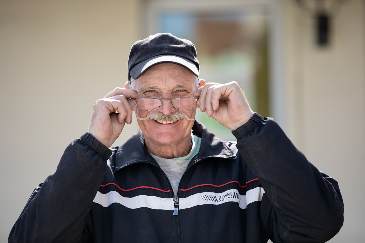 Porträt eines jungen Mannes, der an einen Rezeptions-Tresen lehnt, den rechten Ellbogen darauf aufgestützt. Er lächelt, hat dunkle Haare und trägt einen schwarzen Pullover mit hochgekrämpelten Ärmeln sowie eine helle Bundfaltenhose. In den Händen hält er einen Prospekt.