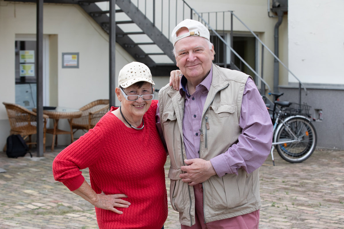 Porträt eines jungen Mannes, der an einen Rezeptions-Tresen lehnt, den rechten Ellbogen darauf aufgestützt. Er lächelt, hat dunkle Haare und trägt einen schwarzen Pullover mit hochgekrämpelten Ärmeln sowie eine helle Bundfaltenhose. In den Händen hält er einen Prospekt.