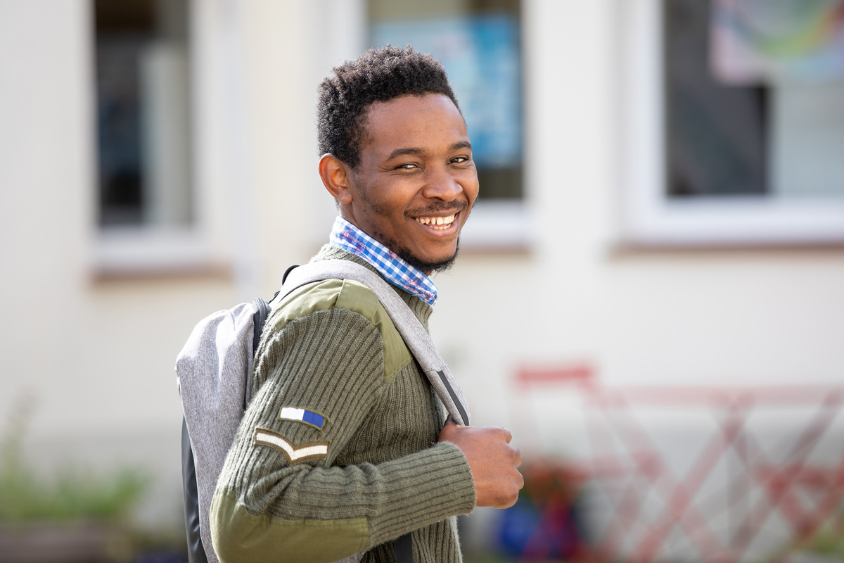 Porträt eines jungen Mannes, der an einen Rezeptions-Tresen lehnt, den rechten Ellbogen darauf aufgestützt. Er lächelt, hat dunkle Haare und trägt einen schwarzen Pullover mit hochgekrämpelten Ärmeln sowie eine helle Bundfaltenhose. In den Händen hält er einen Prospekt.