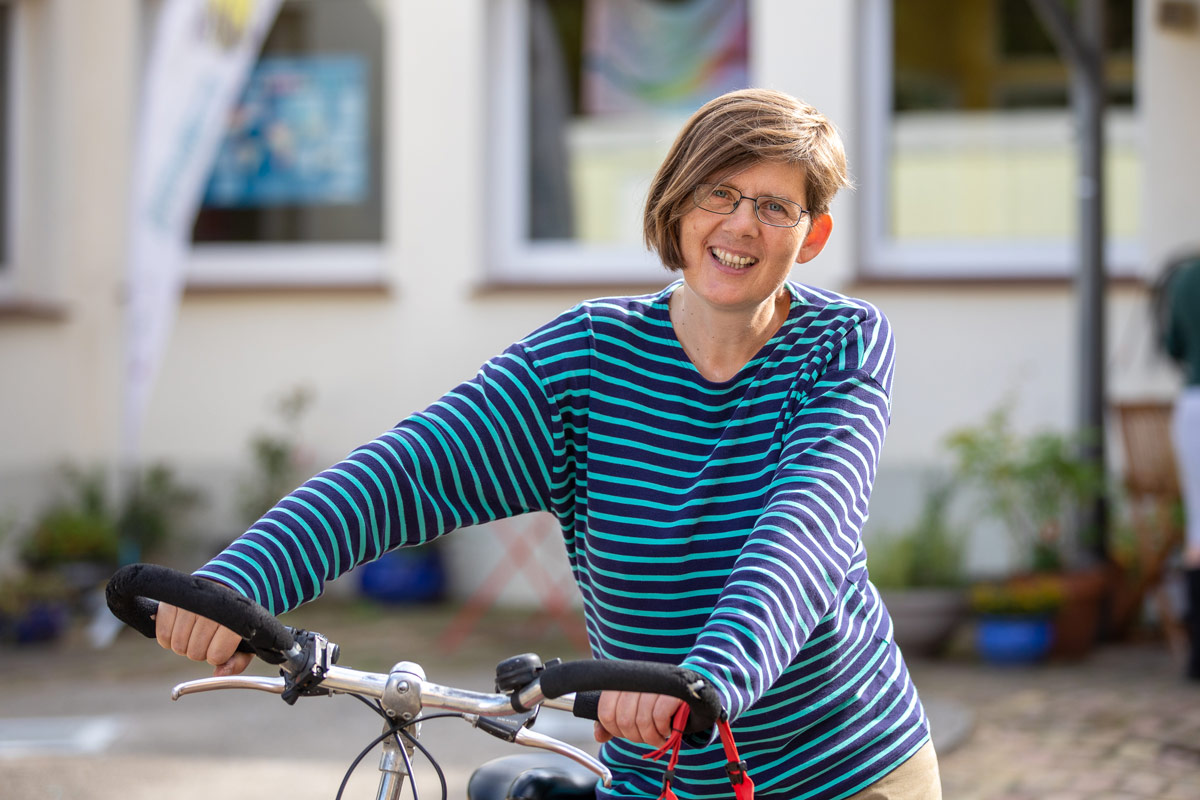 Porträt eines jungen Mannes, der an einen Rezeptions-Tresen lehnt, den rechten Ellbogen darauf aufgestützt. Er lächelt, hat dunkle Haare und trägt einen schwarzen Pullover mit hochgekrämpelten Ärmeln sowie eine helle Bundfaltenhose. In den Händen hält er einen Prospekt.