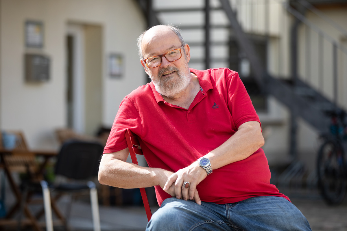 Porträt eines jungen Mannes, der an einen Rezeptions-Tresen lehnt, den rechten Ellbogen darauf aufgestützt. Er lächelt, hat dunkle Haare und trägt einen schwarzen Pullover mit hochgekrämpelten Ärmeln sowie eine helle Bundfaltenhose. In den Händen hält er einen Prospekt.