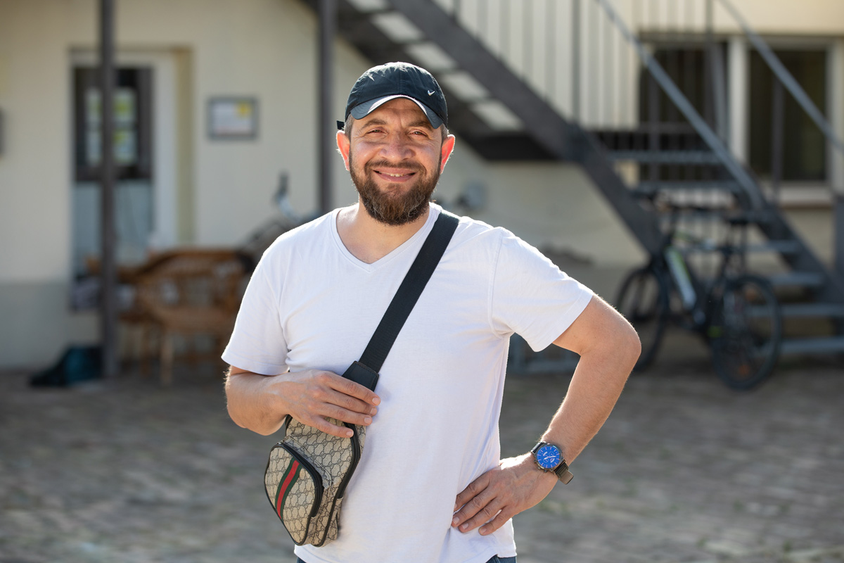 Porträt eines jungen Mannes, der an einen Rezeptions-Tresen lehnt, den rechten Ellbogen darauf aufgestützt. Er lächelt, hat dunkle Haare und trägt einen schwarzen Pullover mit hochgekrämpelten Ärmeln sowie eine helle Bundfaltenhose. In den Händen hält er einen Prospekt.