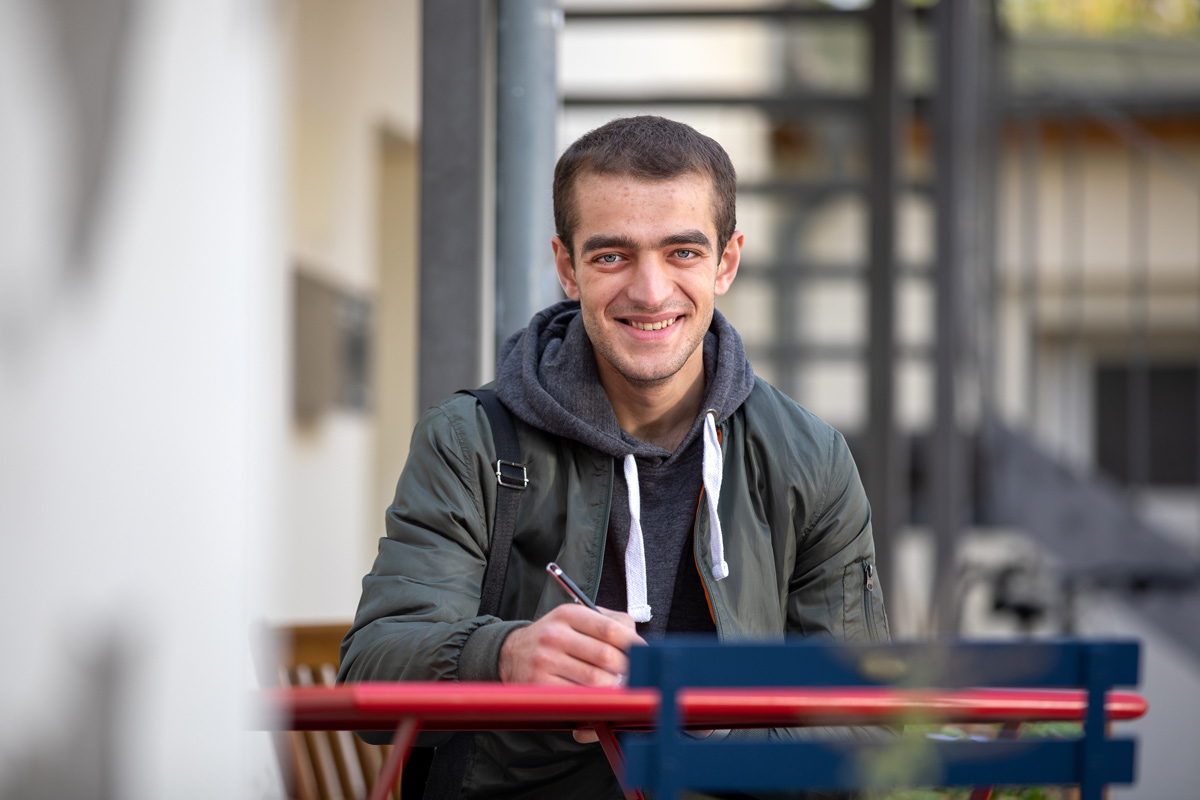 Porträt eines jungen Mannes, der an einen Rezeptions-Tresen lehnt, den rechten Ellbogen darauf aufgestützt. Er lächelt, hat dunkle Haare und trägt einen schwarzen Pullover mit hochgekrämpelten Ärmeln sowie eine helle Bundfaltenhose. In den Händen hält er einen Prospekt.