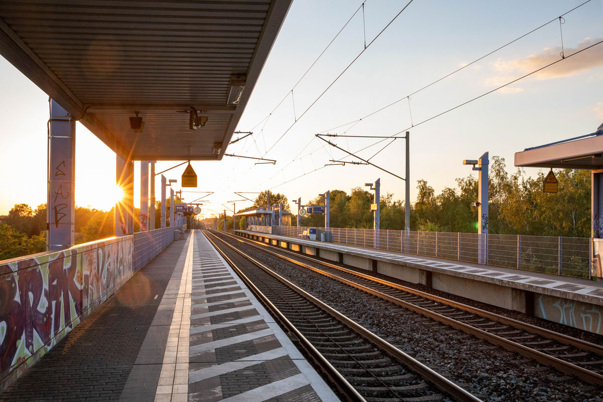 Porträt eines jungen Mannes, der an einen Rezeptions-Tresen lehnt, den rechten Ellbogen darauf aufgestützt. Er lächelt, hat dunkle Haare und trägt einen schwarzen Pullover mit hochgekrämpelten Ärmeln sowie eine helle Bundfaltenhose. In den Händen hält er einen Prospekt.