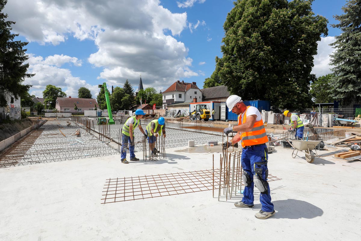 Ein junger dunkelhaariger Mann steht hinter einem Tresen, vor ihm liegt ein Stadtplan. Er lächelt und deutet mit einem Stift auf die Mitte des Planes und erteilt damit einer Frau Auskunft, die sich rechts im Vordergrund des Bildes befindet und ebenfalls auf den Plan blickt.