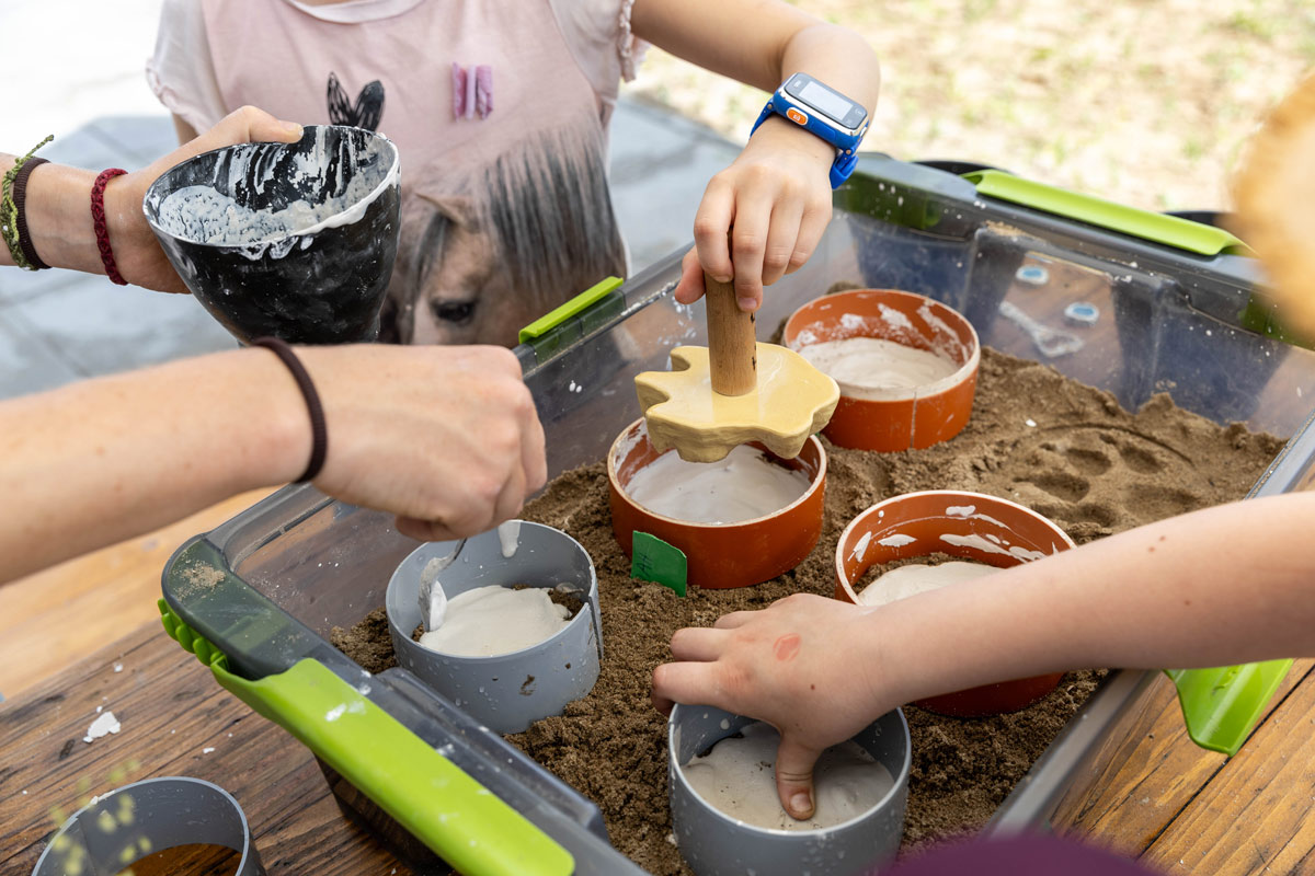 Porträt eines jungen Mannes, der an einen Rezeptions-Tresen lehnt, den rechten Ellbogen darauf aufgestützt. Er lächelt, hat dunkle Haare und trägt einen schwarzen Pullover mit hochgekrämpelten Ärmeln sowie eine helle Bundfaltenhose. In den Händen hält er einen Prospekt.