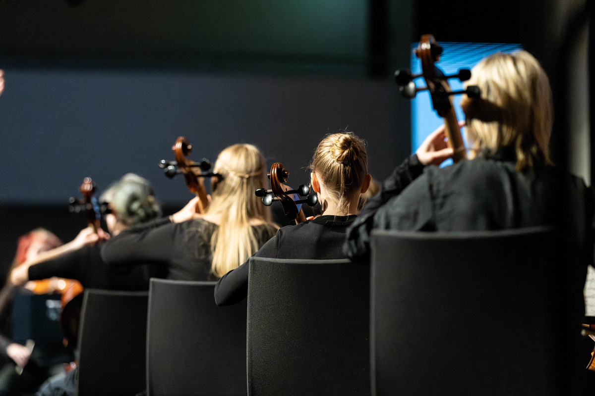 Porträt eines jungen Mannes, der an einen Rezeptions-Tresen lehnt, den rechten Ellbogen darauf aufgestützt. Er lächelt, hat dunkle Haare und trägt einen schwarzen Pullover mit hochgekrämpelten Ärmeln sowie eine helle Bundfaltenhose. In den Händen hält er einen Prospekt.